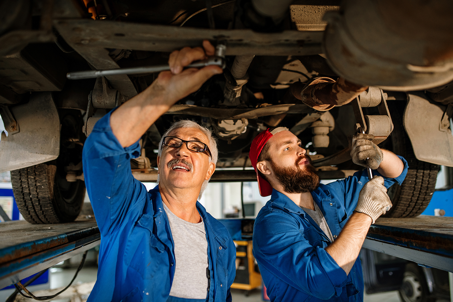 Young and mature technicians of modern car repair service fixing details of car motor while standing under machine